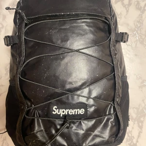 Svart ryggsäck från Supreme - Snygg svart ryggsäck från Supreme i slitstarkt material med dragkedja och justerbara axelremmar. Perfekt för vardagsbruk med stilren design och Supreme-logga i vitt.