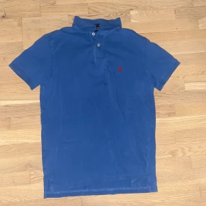 Blå pikétröja från Ralph Lauren - Snygg blå pikétröja från Ralph Lauren med klassisk krage och knappar. Den har en broderad röd logotyp på bröstet. Perfekt för en stilren och avslappnad look. Liten i stoleken cirka 165-175