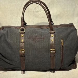  väska från DuffyDuck - Snygg och rymlig väska från DuffyDuck i svart tvättat canvas med bruna skinnhandtag och detaljer. Väskan har en dragkedja upptill och en praktisk ytterficka med dragkedja. Perfekt för resor eller som en stilren vardagsväska. Har även bra fack inuti så du kan ha koll på dina grejer 