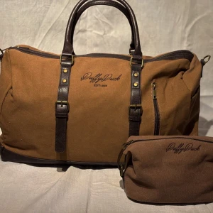Brun weekendväska och necessär från Duffy Duck - Snygg brun weekendväska i canvas med matchande necessär från Duffy Duck. Väskan har mörkbruna skinnhandtag och detaljer, samt en praktisk dragkedja. Perfekt för kortare resor eller som gymväska.