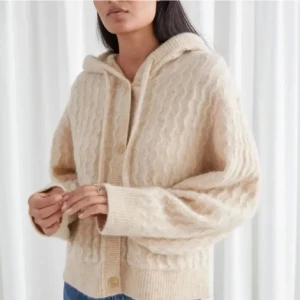 Beige stickad kofta med huva - Mysig beige stickad kofta med huva och knappar framtill. Den har ett vågigt mönster och praktiska fickor. 