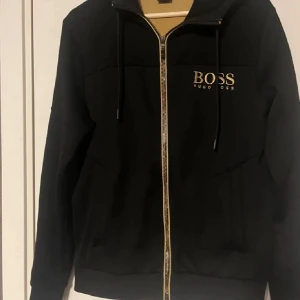 Hugo Boss Zip (Sällsynt) - Snygg svart hoodie från Hugo Boss med dragkedja och huva. Den har märkets logga i guld på bröstet och detaljer på ärmarna. Perfekt för en stilren och bekväm look. Sällsynt modell (SOLBLEKT PÅ LUVAN)