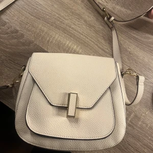 Vit axelväska med gulddetaljer - Snygg vit axelväska i skinn med gulddetaljer. Väskan har en stilren design med ett lock och en praktisk innerficka med dragkedja. Perfekt för att bära dina nödvändigheter med stil.