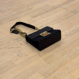 Svart axelväska med gulddetaljer - Snygg svart axelväska med guldkedja och låsdetalj. Väskan har ett elegant mönster och är perfekt för att ge en stilren touch till din outfit. Den kompakta designen gör den lätt att bära med sig. Endast: 249