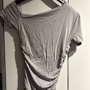 Grå offshoulder topp - Säljer en stilren grå offshoulder topp med korta ärmar. Toppen har en snygg rynkad detalj på sidan som ger en unik look. Storleken är M men passar S perfekt 