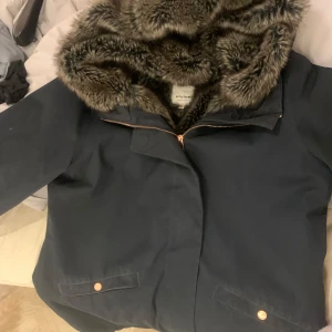 Mörkblå jacka med fuskpäls från Elvine - Säljer en snygg mörkblå jacka från Elvine med mjukt foder i fuskpäls. Jackan har en dragkedja framtill och en stor huva med pälsdetaljer. Perfekt för kyliga dagar när du vill hålla dig varm och stilren. Storlek XL. Priset kan såklart diskuteras 