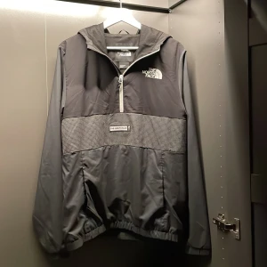 Svart vindjacka från The North Face - Snygg svart vindjacka från The North Face med halv dragkedja och huva. Jackan har ett diskret rutmönster på bröstet och elastiska muddar vid ärmslut och nederkant. Perfekt för blåsiga dagar.