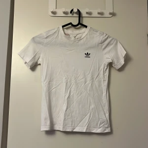Vit t-shirt från Adidas - Säljer en klassisk vit t-shirt från Adidas med deras ikoniska logga i svart på bröstet. T-shirten är kortärmad och tillverkad i bomull, perfekt för en sportig look.