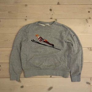 Moncler Sweatshirt - Snygg grå Moncler tröja med ett broderat motiv av en skidåkare på framsidan. Tröjan är extremt tjock och är därför perfekt för afterski. Har en liten slitning vid kragen, men inget som syns särkilt mycket. 