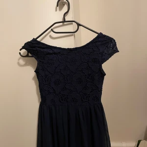 Mörkblå spetsklänning från Cubus - Elegant mörkblå klänning från Cubus med spetsdetaljer och offshoulder-design. Klänningen har en dragkedja i ryggen och en flödande kjol. Perfekt för speciella tillfällen.