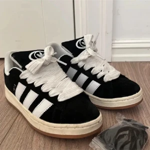 Svarta Adidas Campus sneakers - Snygga svarta Adidas Campus sneakers med vita ränder och breda snören. Skorna har en klassisk design med gummisula och är perfekta för en avslappnad stil.
