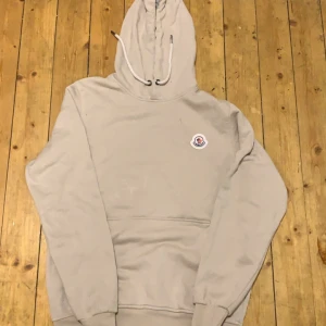 Beige hoodie från Moncler - Säljer en stilren beige hoodie från Moncler med justerbar huva och ribbade muddar. Perfekt för en avslappnad look. Moncler-logga på bröstet ger en exklusiv touch.