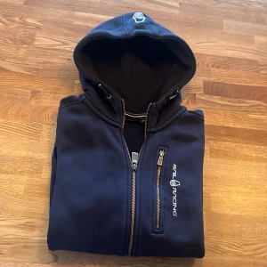 Mörkblå hoodie från Sail Racing - Säljer en mörkblå hoodie från Sail Racing med dragkedja och logotyp på bröstet. Perfekt för kyliga dagar med sin bekväma passform och huva. Använd väldigt få gånger då den var för liten från början. Strl 160. Köpt på kids brandstore, nypris 899 kvitto följer med. 