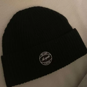 Svart ribbad mössa från Axel Arigato - Snygg svart ribbad mössa från Axel Arigato med en stilren broderad logga framtill. Perfekt för att hålla värmen under kyliga dagar. Använd få tal gånger, säljs inte längre på marknaden. Helt ny.