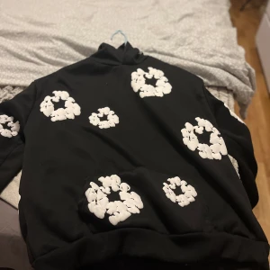 Svart hoodie med vita blommor från Denim Tears - Snygg svart hoodie från Denim Tears med vita blommönster över hela tröjan. Perfekt för en unik och stilren look. Hoodien har en klassisk passform med långa ärmar och en bekväm huva.