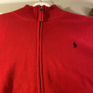 Röd tröja med dragkedja från Polo Ralph Lauren - Snygg röd tröja från Polo Ralph Lauren med dragkedja och ribbad krage. Tröjan har långa ärmar med svarta ränder och en broderad logotyp på bröstet. Perfekt för en stilren look.