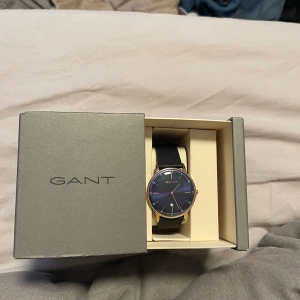 Gant Phoenix GT046005 - Elegant klocka från Gant med en stilren blå urtavla och guldfärgad boett. Klockan har ett svart läderarmband som ger en klassisk look. Perfekt för den som vill ha en tidlös accessoar.