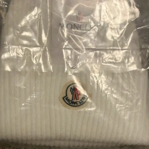 Vit ribbad mössa från Moncler - Snygg vit ribbad mössa från Moncler med broderad logotyp framtill. Perfekt för att hålla värmen under kyliga dagar. Köpte den begagnat så vet it om den är äkta.