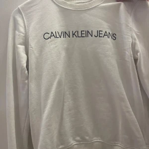 Vit sweatshirt från Calvin Klein Jeans - Snygg vit sweatshirt från Calvin Klein Jeans med svart logotyptryck på bröstet. Tröjan har långa ärmar och en rund halsringning. Perfekt för en stilren och avslappnad look.