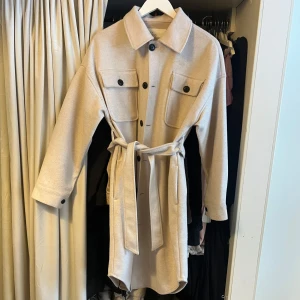 Beige kappa från NLY Trend - Snygg beige kappa från NLY Trend med knappar och knytskärp i midjan. Jackan har långa ärmar och två stora bröstfickor med knappar. Perfekt för en stilren look.