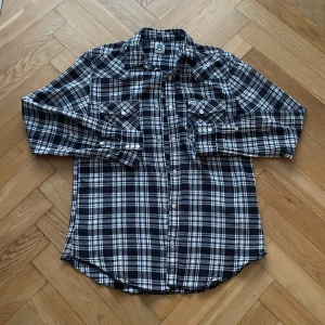 Svart och vit rutig skjorta från Wrangler - Snygg svart och vit rutig skjorta från Wrangler med långa ärmar och klassisk krage. Skjortan har knappar framtill och två bröstfickor med knappstängning. Perfekt för en avslappnad stil.