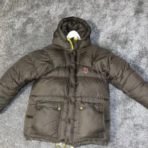 Grön dunjacka från Fjällräven - Säljer en grön dunjacka från Fjällräven med huva och gula detaljer. Jackan har en dragkedja och knappar framtill samt praktiska fickor. Perfekt för kyliga dagar.