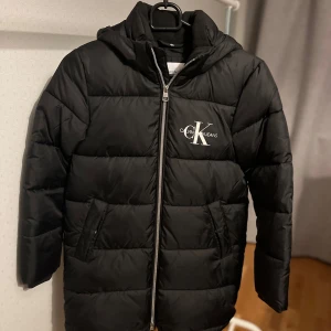 Svart pufferjacka från Calvin Klein - Snygg svart pufferjacka från Calvin Klein med dragkedja och huva. Jackan har en stilren design med CK-logga på bröstet och praktiska fickor. Perfekt för kyliga dagar. Priset kan diskuteras, vid frågor är det bara att höra av sig👍🏼👍🏼