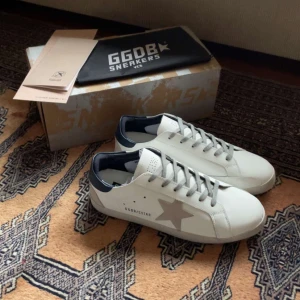 Vita sneakers från Golden Goose - Snygga vita sneakers från Golden Goose med grå snörning och en stjärndetalj på sidan. Skorna har en kontrasterande blå häl och en bekväm sula. Perfekta för en stilren look.