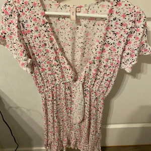 Blommig klänning från Nelly - Söt blommig klänning från Nelly i vitt med rosa och svarta blommor. Klänningen har en v-ringad design och korta ärmar. Perfekt för en somrig look med knytning i midjan för en smickrande passform. Passar perfekt till midsommar, klänningen har använts en gång och har inga defekter