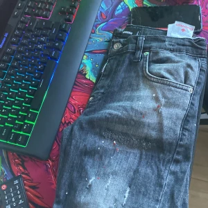 Svarta dsquared2 jeans ganska nya  - Snygga svarta jeans från Dsquared2 med unika färgstänk och slitningar. De har en klassisk femficksdesign och knappgylf. Perfekta för en trendig och avslappnad stil.
