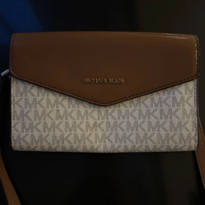Brun och beige axelväska från Michael Kors - Snygg axelväska från Michael Kors i brunt och beige med logotypmönster. Väskan har en elegant design med en brun läderdetalj på locket och en justerbar axelrem. Fack i baksidan! Skriv för mer info eller bilder❤️