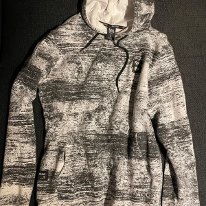Svartvit mönstrad hoodie Black Squad - Snygg svartvit hoodie, Black Squad. Tröjan har en justerbar huva och en praktisk magficka. 