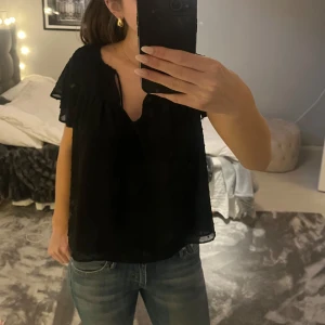 Jätte fin svart blus💕 - Snygg svart blus med korta ärmar och volangdetaljer. Blusen har ett diskret prickigt mönster och en v-ringad hals. Perfekt för en stilren look.