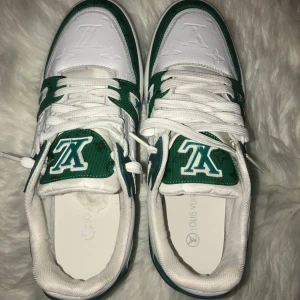 Vita och gröna sneakers från Louis Vuitton - Snygga vita och gröna sneakers från Louis Vuitton med märkets logga på tungan och hälen. Skorna har en stilren design med snörning och en bekväm sula. Perfekta för en trendig look.