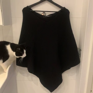 Svart stickad poncho - Säljer en stilren svart stickad poncho som är perfekt för kyliga dagar. Den har en asymmetrisk design och är lätt att slänga över en tröja för extra värme. Passar bra till både vardag och lite finare tillfällen.