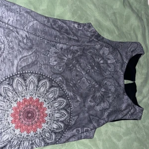 Grå ärmlös klänning - Snygg grå ärmlös klänning med detaljerat mandalamönster i vitt och rött. Klänningen har en rund hals och en liten öppning bak i nacken. 