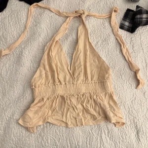 Beige halterneck topp - Snygg beige halterneck topp med knytband i nacken och smockdetaljer i midjan. Toppen har en lätt volangkant nedtill och är perfekt för varma dagar.
