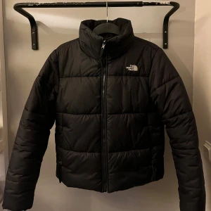 Svart pufferjacka från The North Face - Säljer en svart pufferjacka från The North Face (Dam). Jackan har en dragkedja framtill och är perfekt för kyligare väder. Den har en hög krage för extra värme och stil. Jackan har även innerficka. Inga hål, helt ren.