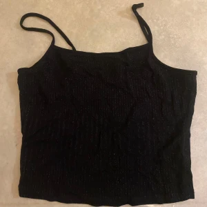 Svart glittrig linnetopp från BikBok - Snygg svart glittrig linnetopp från BikBok med smala axelband. Perfekt för en kväll ute eller en festlig tillställning. Toppen har en glittrig finish som ger en extra touch av glamour.