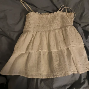 Vit topp från Gina Tricot - Söt vit topp från Gina Tricot med smala axelband och smockdetaljer upptill. Toppen har ett volangliknande fall och små glittriga detaljer som ger en extra touch. Perfekt för varma sommardagar!