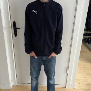 Svart hoodie från Puma - Säljer en marinblå Puma tröja i storlek YXL (passar ca 160–170 cm). Den har en dragkedja framtill, hög krage och Puma-logga tryckt på bröstet. Lätt och bekvämt material som passar för träning eller vardagsbruk.  Personen på bilderna är 160 cm lång, 12 år gammal, och har en smal/normal kroppsbyggnad.  Skick: Bra begagnat skick, med minimala tecken på användning.  Nypris: 400 SEK