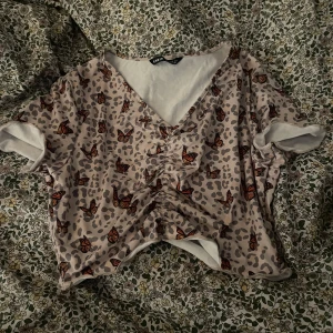 Beige topp med fjärilsmönster från Shein - Säljer en superfin beige topp från Shein med ett coolt fjärilsmönster. Toppen har en v-ringad design och korta ärmar, perfekt för sommardagar. Materialet känns lätt och luftigt, vilket gör den skön att bära. Passar perfekt till både jeans och kjol!