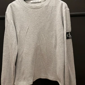 Grå långärmad tröja från Calvin Klein Jeans - Säljer en stilren grå långärmad tröja från Calvin Klein Jeans. Den har en diskret textur och ett snyggt CK-märke på ärmen. Perfekt för en casual look eller att klä upp med accessoarer. Passar till alla säsonger!