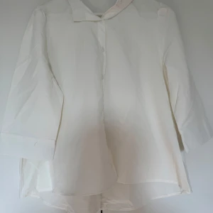 Vit blus med trekvartsärm - Säljer en stilren vit blus med trekvartsärm. Perfekt för både vardag och festliga tillfällen. Blusen har en klassisk krage och knappar framtill. Den är i ett lätt och luftigt material som gör den bekväm att bära hela dagen.