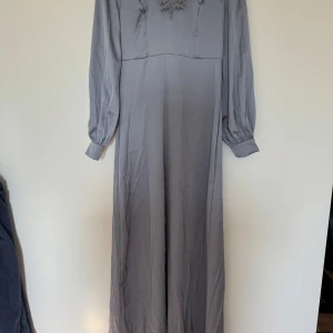 Grå långklänning med spetsdetaljer - Säljer en elegant grå långklänning med vackra spetsdetaljer vid halsringningen. Klänningen har långa ärmar med knappar vid ärmsluten och en mjuk, flytande passform. Perfekt för höst- och vinterfester!