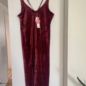 Vinröd sammet klänning - Säljer en elegant vinröd klänning i mjuk sammet. Den har en lång design med smala axelband och en djup v-ringning. Perfekt för festliga tillfällen eller en utekväll. Klänningen är i nyskick och ger en lyxig känsla.