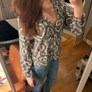Mönstrad blus i svart och beige - Säljer en snygg blus med ett coolt mönster i svart och beige. Blusen har långa ärmar och en v-ringad design som ger en stilren look. Perfekt för både vardag och fest!