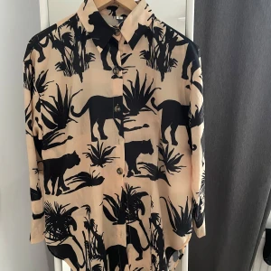 Beige och svart mönstrad skjorta från Shein - Säljer en supercool beige skjorta från Shein med ett unikt svart djur- och växtmönster. Skjortan har långa ärmar och knappar framtill. Perfekt för att sticka ut på festen eller bara ha en chill dag. Den är i storlek S och är i nyskick. Passar perfekt till både jeans och kjol!