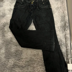 Svarta jeans från River Island - Säljer ett par snygga svart/gråa lågmidjade, bootcut/straight, oanvända jeans från River Island. De har en klassisk femficksdesign med coola dragkedjedetaljer på bakfickorna. Storleken på lappen är en 14, M/L