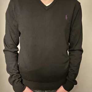 Svart tröja från Ralph Lauren - Storlek m, Säljer en stilren svart tröja från Ralph Lauren i merinoull. Tröjan har en v-ringad design och ett lila broderat logotypmärke på bröstet. Perfekt för en elegant och bekväm look.